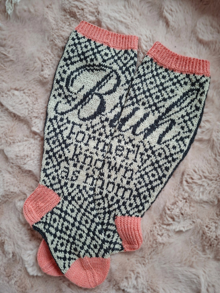 Knitted socks with a pattern and pink accents and text that reads "Bruh Formerly known as mom" on a textured surface