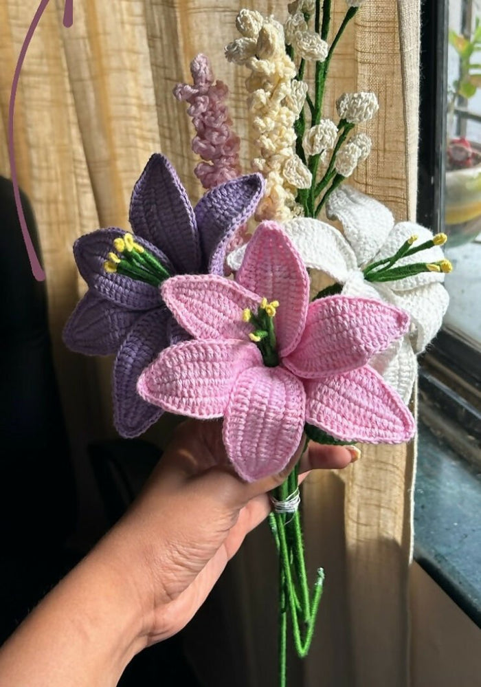 Hand holding a bouquet of pink, purple, and white crocheted flowers against a neutral background.