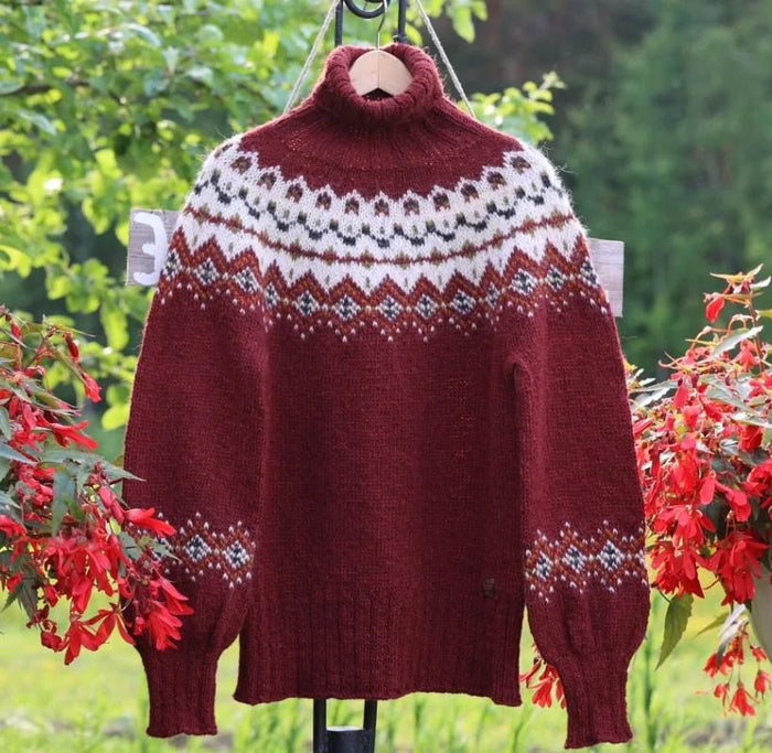 A burgundy knit turtleneck sweater with a white, gray, and black stranded colorwork circular yoke pattern, hanging on a wooden hanger outdoors against a green garden background with red flowers.
