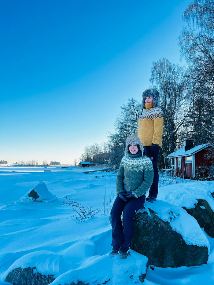 Two people posing in a snowy outdoor winter landscape under a clear blue sky. One person is seated on a snow-covered rock wearing a gray knit sweater, and the other stands behind them wearing a yellow knit sweater with a patterned collar and a gray fur-trimmed hat.