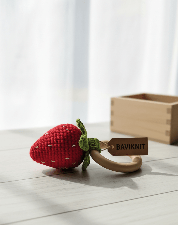 Red strawberry-shaped teething ring with a wooden tag labeled 'BAVIKNIT' on a light wooden surface.