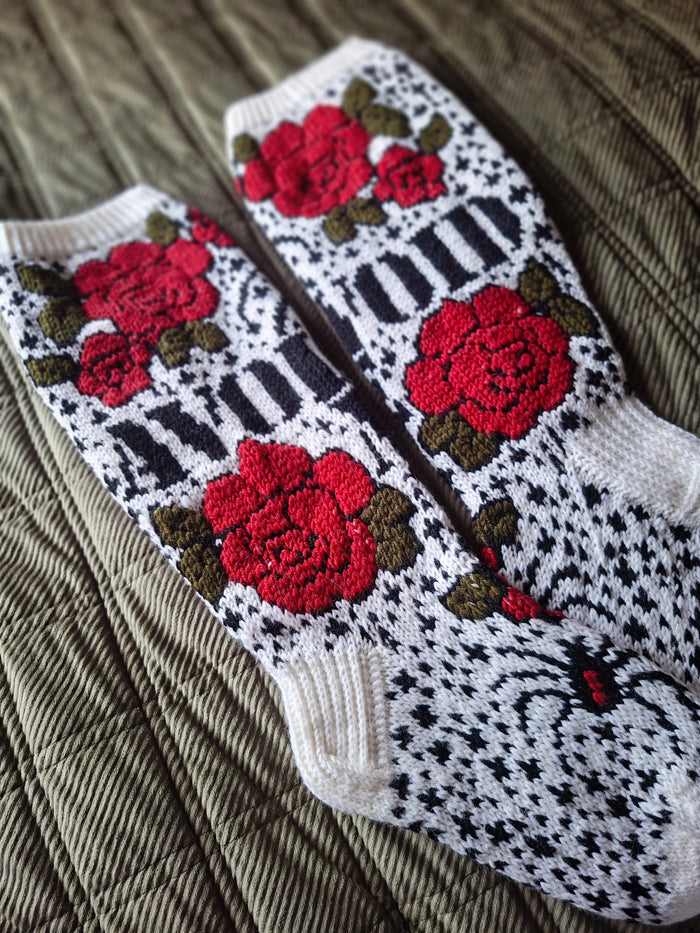 A pair of hand-knit socks featuring red roses with green leaves, black and white stripes, and a black spider motif on a white background, laid flat on a quilted surface.