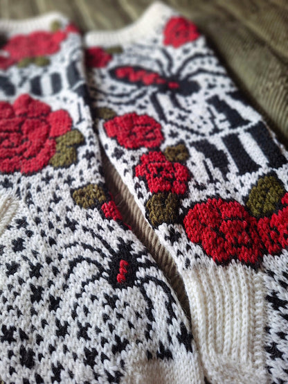 Close-up of a pair of hand-knit socks featuring a stranded colorwork pattern with red roses, black and white geometric motifs, and olive green leaves.
