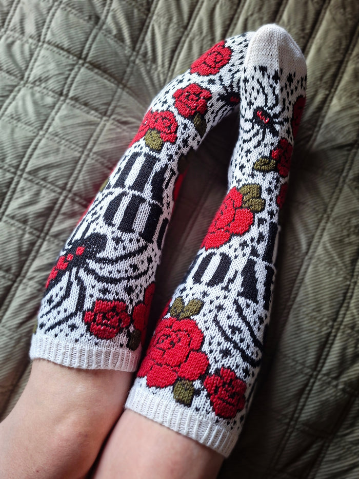 A pair of white knit socks with a black geometric pattern and red rose embroidery, worn on feet resting on a quilted blanket.
