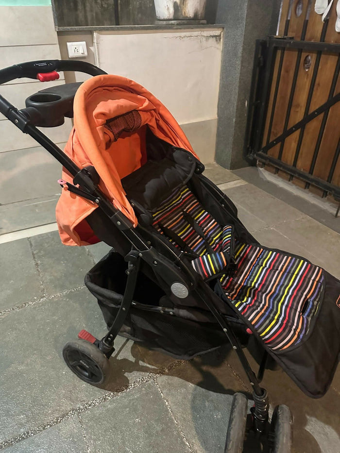 Black stroller with orange cover and colorful striped footmuff on a stone pavement.