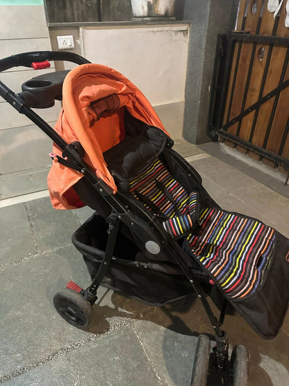 Black stroller with orange cover and colorful striped footmuff on a stone pavement.