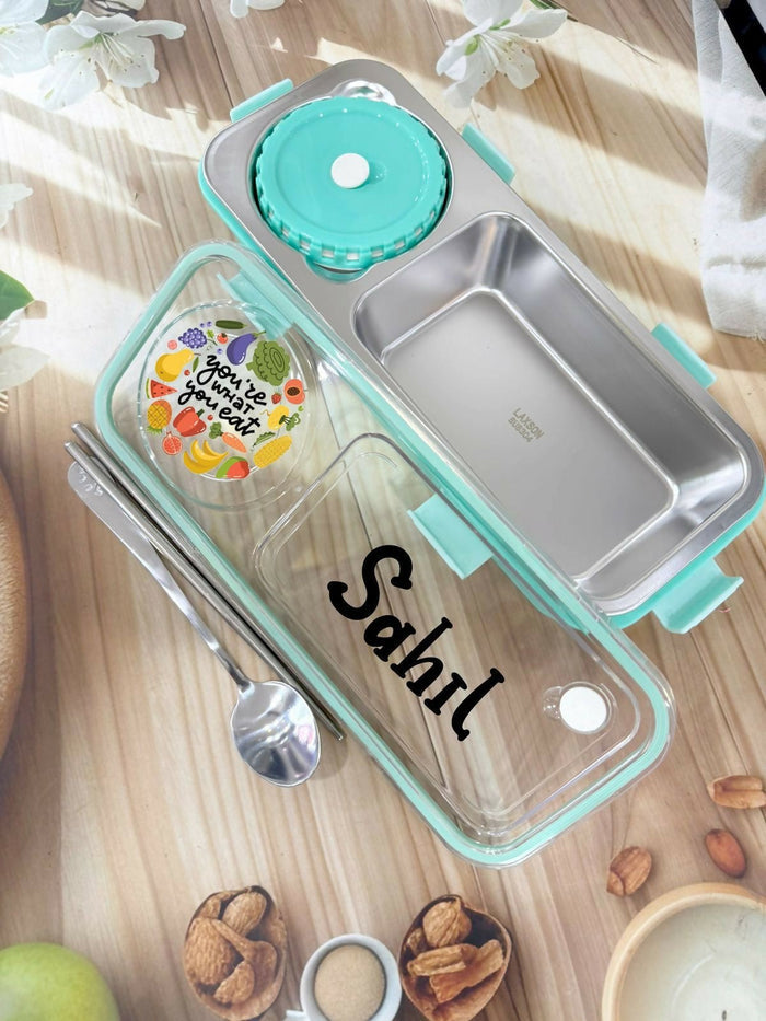 Stainless steel bento box with clear compartment, turquoise lid, and personalized name 'Sahil' on a wooden surface.