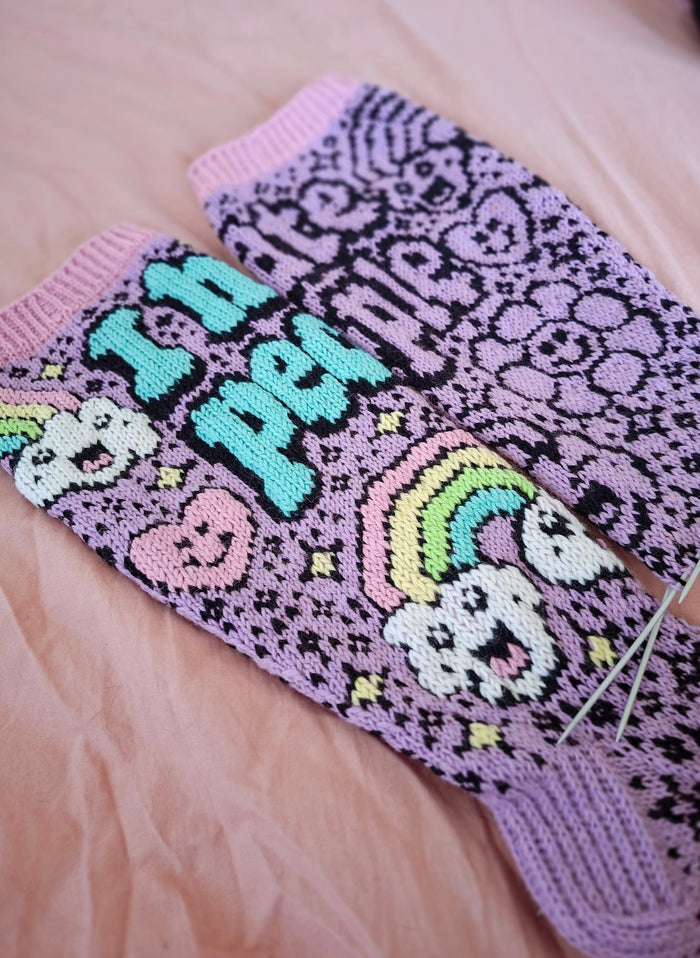 Knitted socks with text and patterns on a pink fabric background. One sock has colorful duplicate stitching the second does not