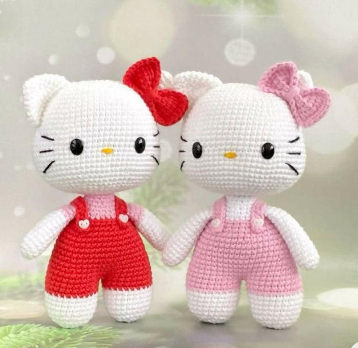 Two crochet Hello Kitty dolls, one in red overalls and the other in pink, against a blurred background.