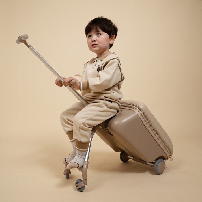 Child sitting on a suitcase with a rolling toy, both in beige, against a beige background