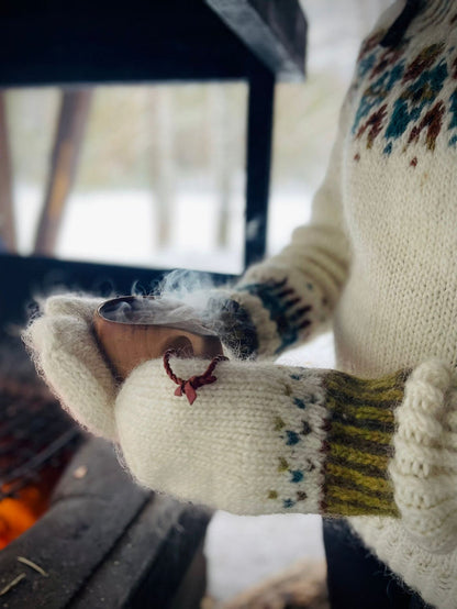 A close-up shot of a person wearing a cream-colored knit sweater and mittens with a stranded colorwork pattern, holding a kuksa over a dark fireplace in a winter setting.
