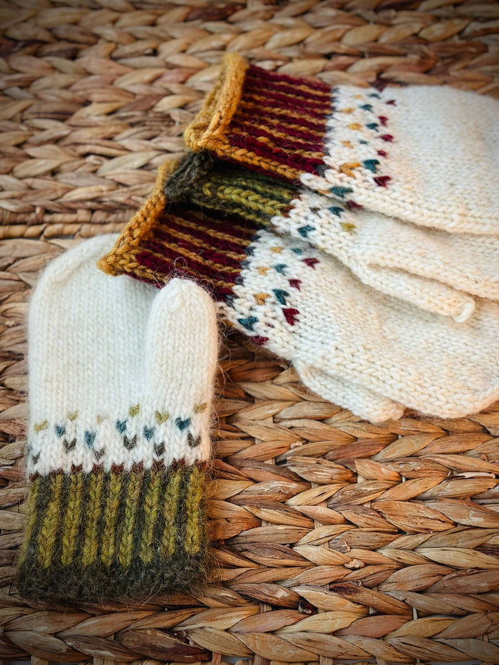 Hand-knitted lightly felted mittenss in cream wool with corrugated ribbing and a stranded colorwork patterns, displayed on a woven straw mat.