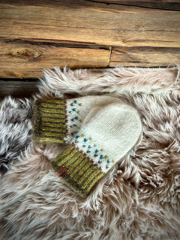 A pair of hand-knit wool mittens in cream, olive green, and blue, styled on a fluffy faux fur rug with a rustic wooden plank background.