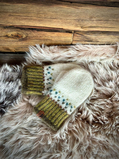 A pair of hand-knit wool mittens in cream, olive green, and blue, styled on a fluffy faux fur rug with a rustic wooden plank background.