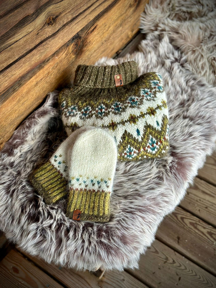 A knit sweater and matching mittens laid on a shaggy faux-fur rug over a wooden floor. The sweater is olive green and cream with a stranded colorwork pattern in blue, red, and yellow, and the mittens are cream with olive green and blue accents.