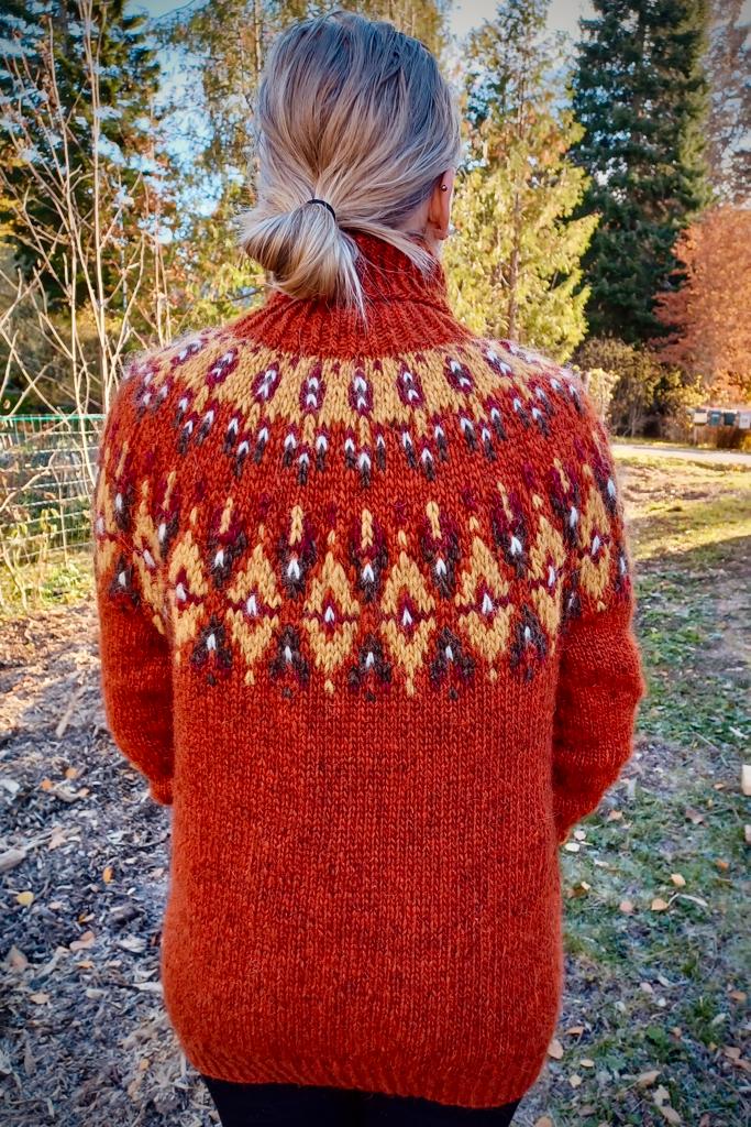A person viewed from behind wearing a knit turtleneck sweater outdoors. The sweater has a warm rust-orange base with a largestranded colorwork circular yoke pattern in cream, brown, and dark red accents.