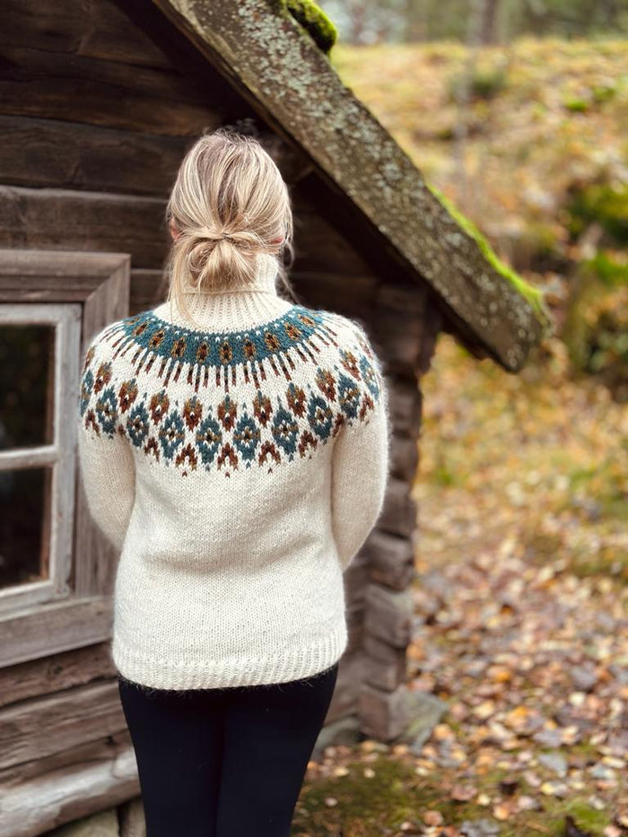 A woman viewed from behind wearing a cream-colored knit sweater with a detailed stranded colorwork circular yoke pattern, standing in front of a rustic wooden cabin outdoors.