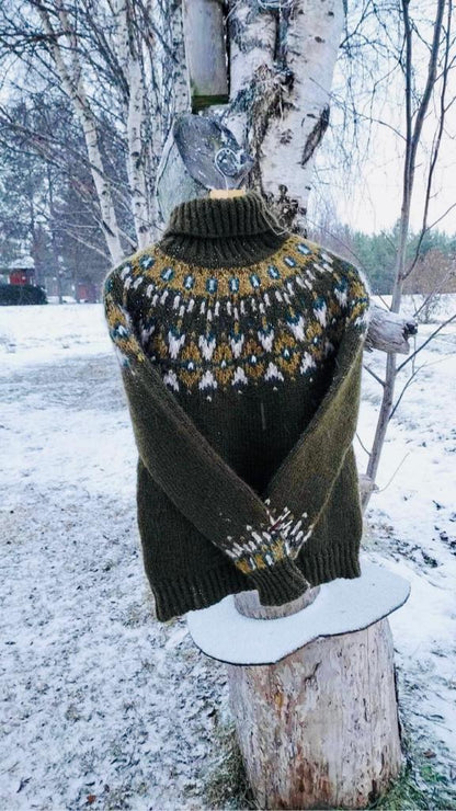 An olive green knit turtleneck sweater with a colorful stranded colorwork circular yoke pattern, displayed hanging off a tree outdoors in a snowy forest setting.