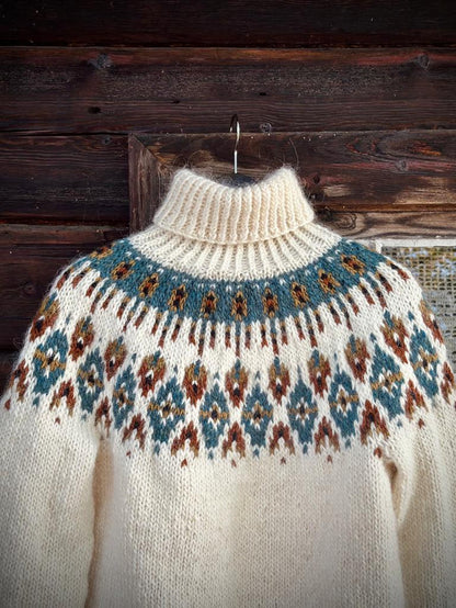 A cream-colored knit turtleneck sweater with a blue, brown, and orange stranded colorwork circular yoke pattern, hanging on a wooden hanger against a rustic dark wood plank wall.