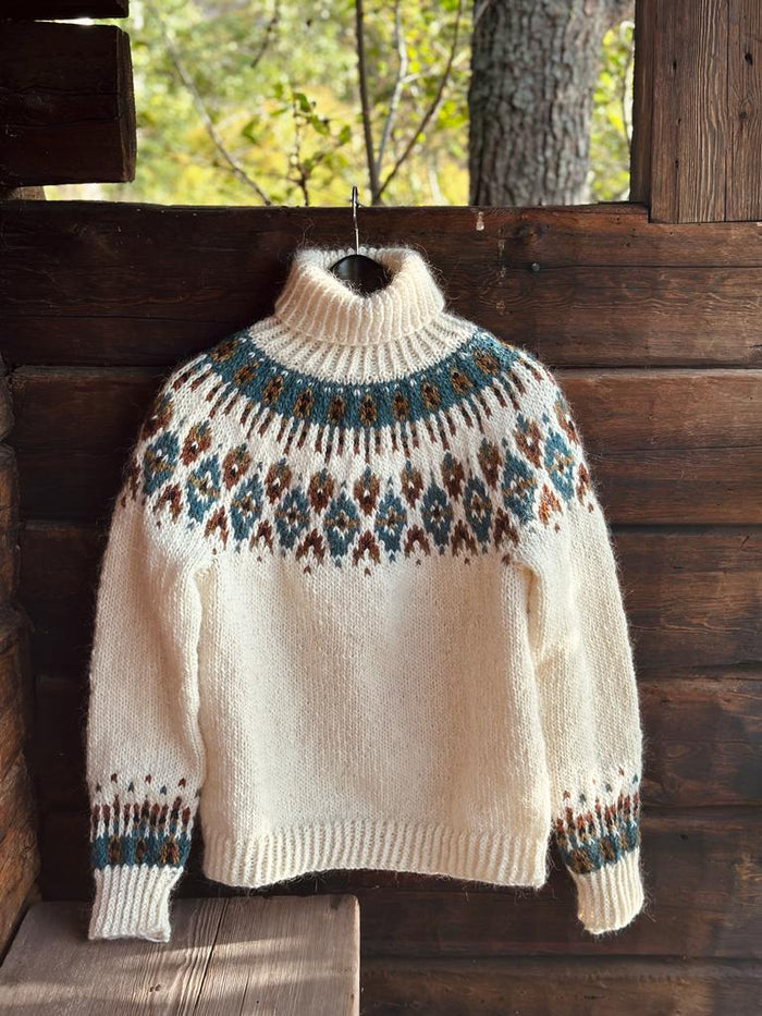 A cream-colored knit turtleneck sweater with a blue and brown stranded colorwork circular yoke pattern, hanging on a wooden porch.