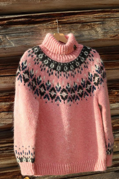 A pink knit turtleneck sweater with a black and white stranded colorwork circular yoke pattern, hanging on a wooden hanger against a rustic wooden wall.