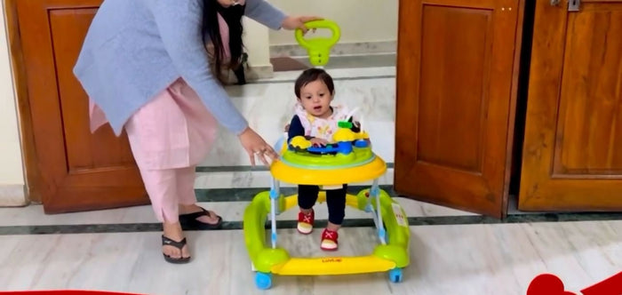 Child using a yellow and green walker with a person assisting, indoors.