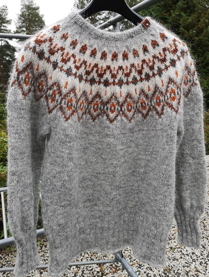 A gray hand-knit sweater with a brown and rust orange stranded colorwork circular yoke pattern, hanging on a black hanger outdoors.