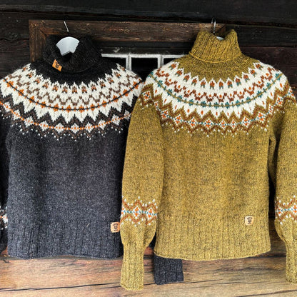 Two hand-knit turtleneck sweaters with the same stranded colorwork circular yoke pattern hanging on a rustic wooden surface. The sweater on the left is dark charcoal gray with a black, white, and orange pattern. The sweater on the right is a mustard yellow with white, orange, and teal.