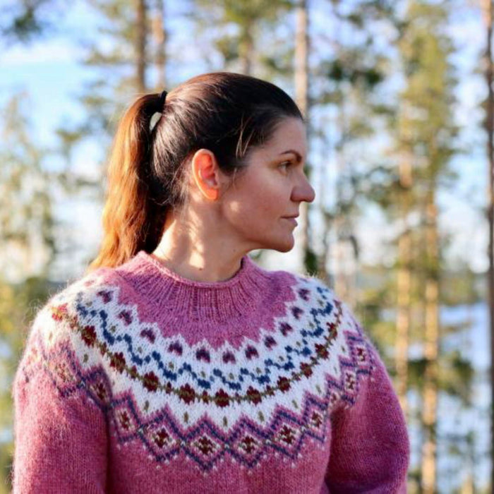 A woman in profile wearing a pink stranded colorwork circular yoke knit sweater with white, blue, and brown patterns, set against a blurred background of pine trees and water.