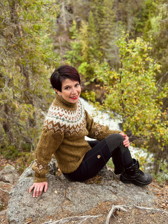 A woman with short dark hair sits on a large gray rock in a forest setting, wearing a brown stranded colorwork knit turtleneck sweater with cream and beige geometric patterns, black pants, and black boots.