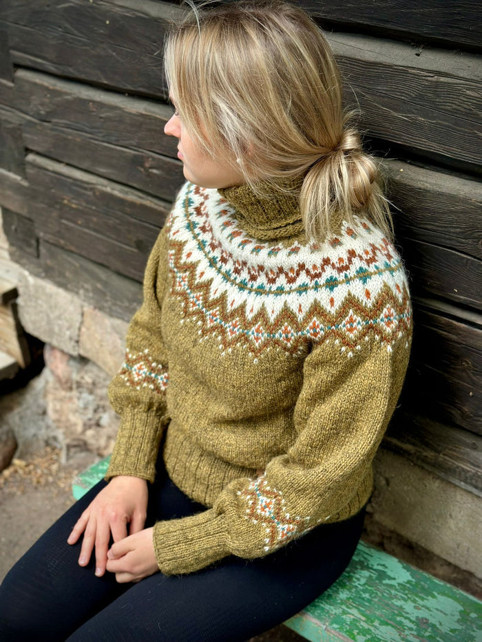 A woman wearing a hand-knit turtleneck sweater with a stranded colorwork pattern in olive green, cream, brown, and teal, sitting against a dark wooden wall.