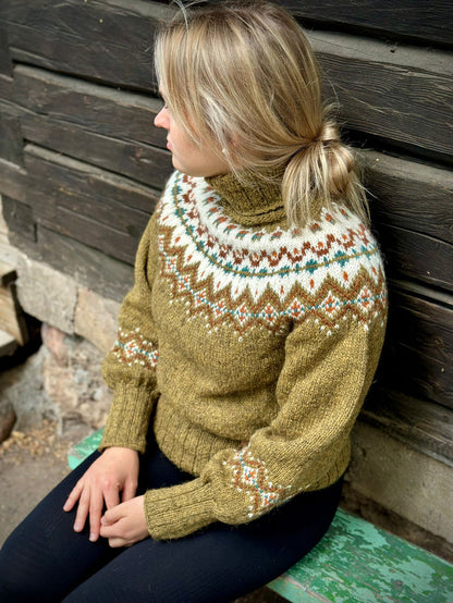 A woman wearing a hand-knit turtleneck sweater with a stranded colorwork pattern in olive green, cream, brown, and teal, sitting against a dark wooden wall.
