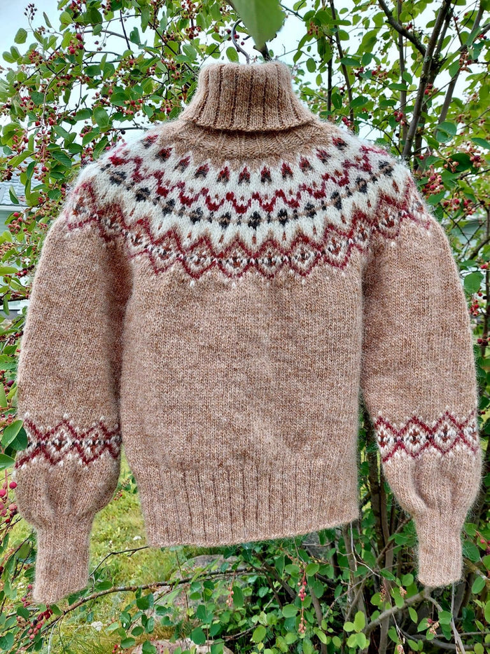 A hand-knit turtleneck sweater in a warm taupe-brown color, featuring a stranded colorwork circular yoke pattern in cream, burgundy, and black, with matching geometric patterns on the cuffs. The sweater is displayed outdoors against a background of green leaves and small red berries.