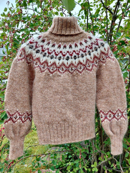 A hand-knit turtleneck sweater in a warm taupe-brown color, featuring a stranded colorwork circular yoke pattern in cream, burgundy, and black, with matching geometric patterns on the cuffs. The sweater is displayed outdoors against a background of green leaves and small red berries.