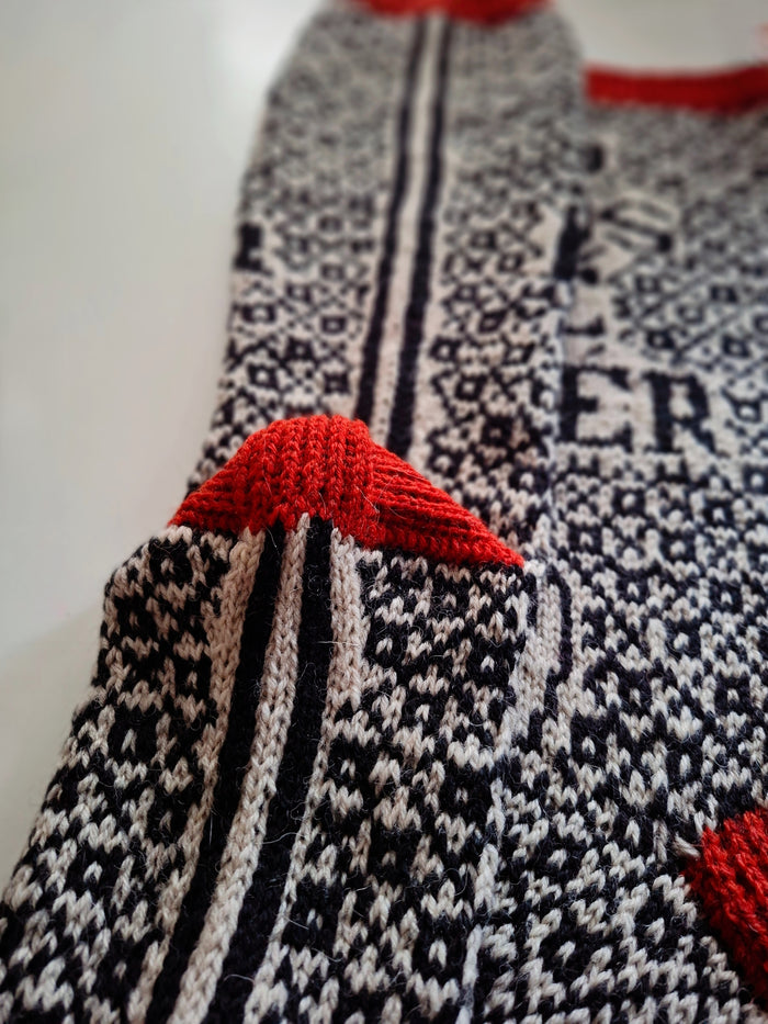 Close up o the heel of a knitted sock with a black and white pattern and red accents on a white background