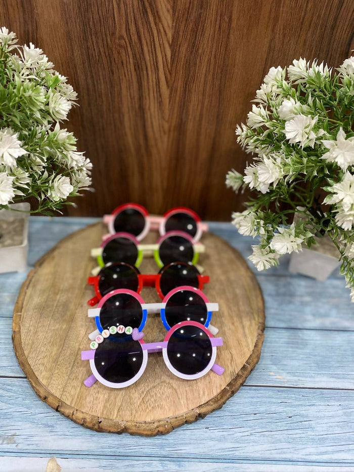 Set of colorful children's sunglasses on a wooden board with flowers on either side.