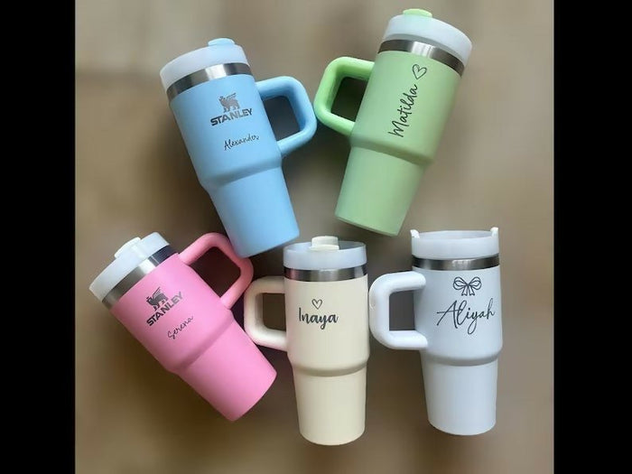 Five insulated travel mugs in different colors with names engraved on them against a brown background.
