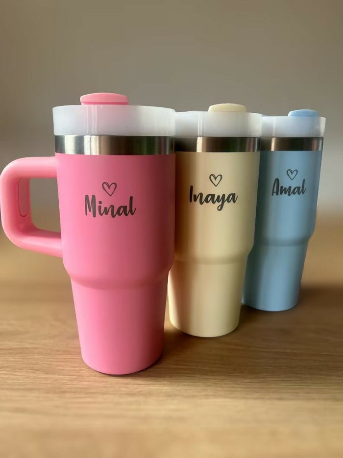 Three insulated travel mugs in pink, beige, and blue with names 'Minal', 'Naya', and 'Amal' on a wooden surface.