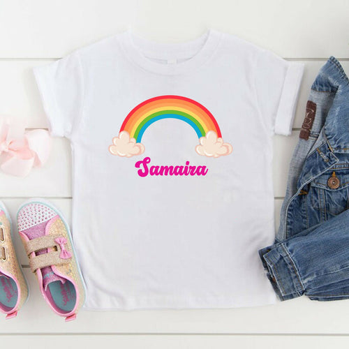 White t-shirt with rainbow and 'Samaira' design, surrounded by children's shoes and denim jacket on a white surface.