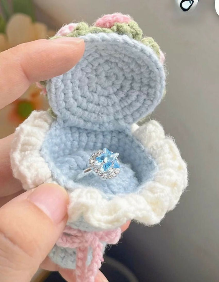 Crocheted ring holder with a blue ring inside, held by a hand against a blurred background.