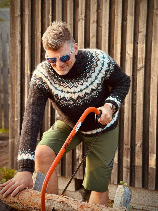 A man wearing a dark gray and cream stranded colorwork patterned knit sweater, olive green shorts, and sunglasses is seen sawing a log outdoors against a wooden fence.