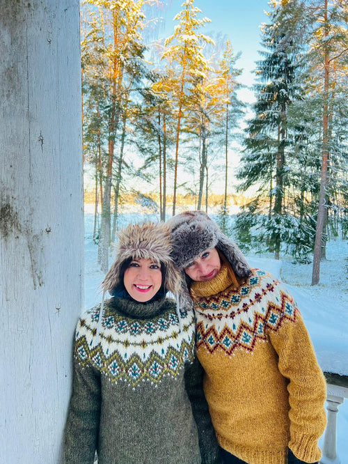 Two people posing outdoors in a snowy forest setting, wearing cozy knit stranded colorwork sweaters and furry ear flap hats. One person wears a dark gray-green sweater with cream, blue, and red geometric patterns, while the other wears a mustard yellow sweater with a white, red, and blue pattern.