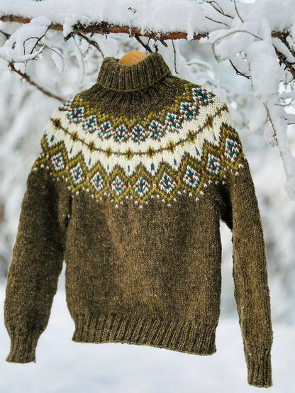 A hand-knit turtleneck sweater in olive green with a colorful stranded colorwork circular yoke pattern, hanging on a wooden hanger from a snow-covered tree branch.