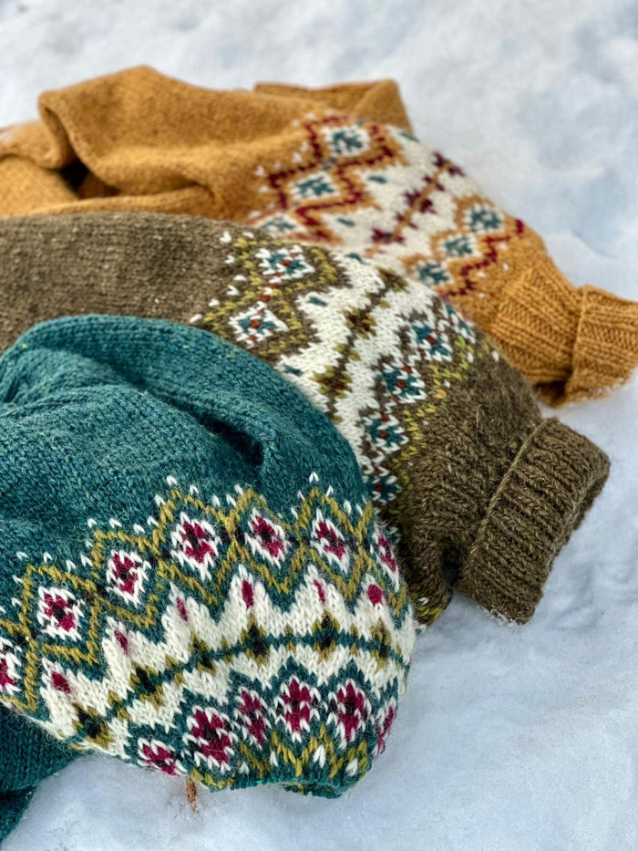 Three knitted sweaters in different colors including teal, mustard, and brown, arranged on a white snowy background.