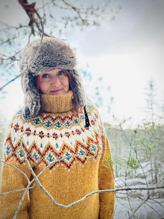 A woman wearing a cozy mustard yellow knit sweater with a colorful stranded colorwork circular yoke pattern, a matching knit turtleneck, and a gray furry hat, standing outdoors in a snowy, wooded setting.