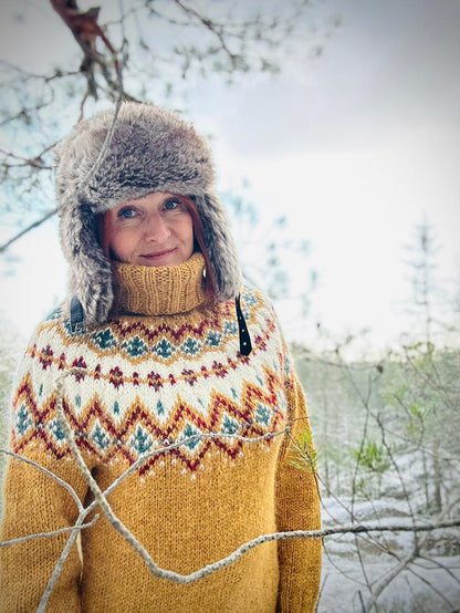 A woman wearing a cozy mustard yellow knit sweater with a colorful stranded colorwork circular yoke pattern, a matching knit turtleneck, and a gray furry hat, standing outdoors in a snowy, wooded setting.