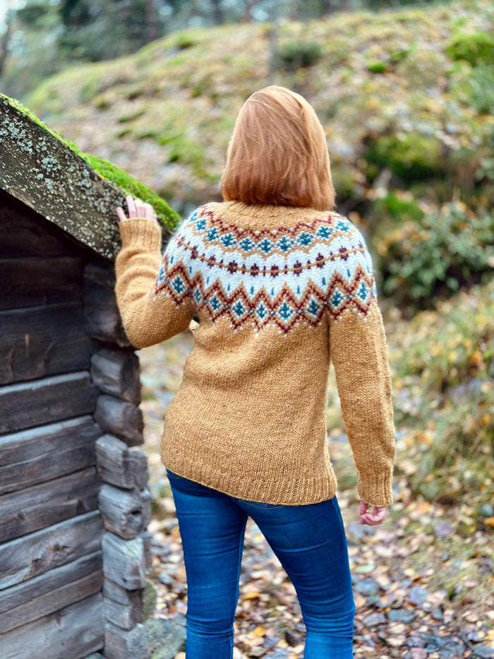 A woman viewed from behind wearing a yellow knit sweater with a colorful stranded colorwork circular yoke pattern in blue, brown, and cream, standing outdoors next to a rustic wooden cabin.