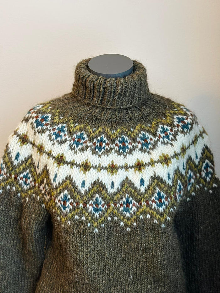 A close-up of a hand-knit turtleneck sweater displayed on a mannequin torso. The sweater features a dark brown base with a prominent white yoke decorated with a stranded colorwork pattern in olive green, blue, and red.