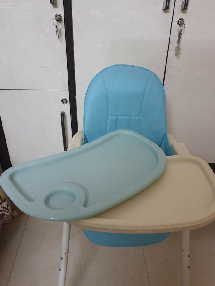 Children's high chair with blue cushion and beige tray against a white wall.