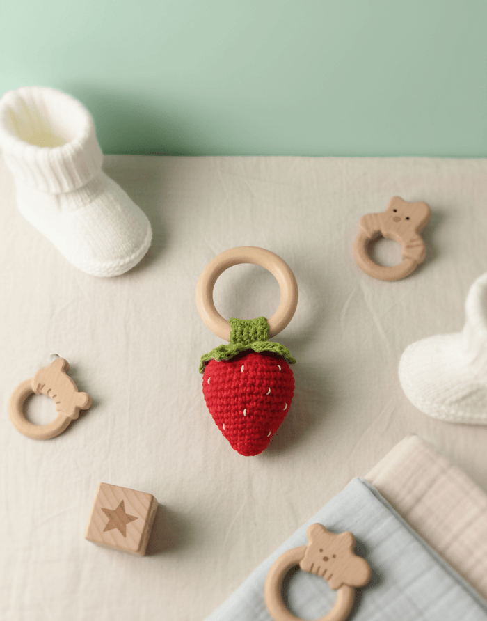 Strawberry-shaped teething ring with wooden rings and baby booties on a light surface.